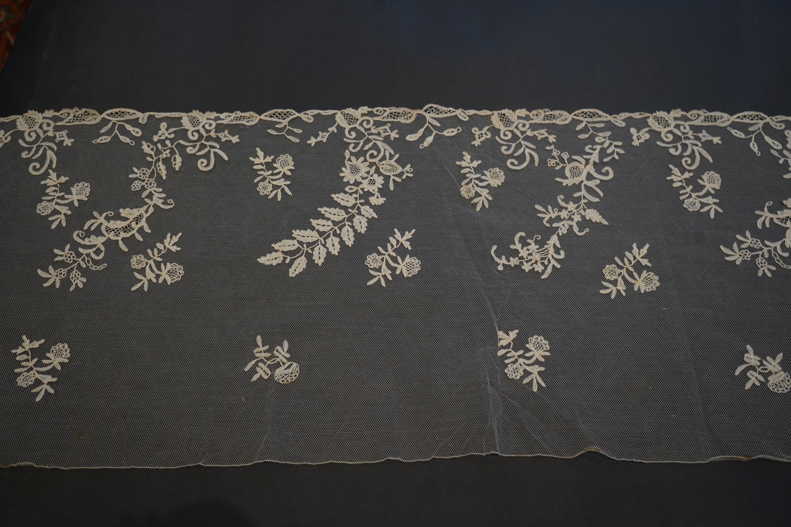 A 19th century five yard flounce of Brussels bobbin appliqué lace applied onto fine machine net, worked in a trailing floral design with an ornate border and sprig motifs, 457.5 cm long x 36cm deep. Condition - possibly